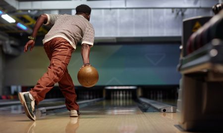 Man performing a bowling release in modern bowling alley with focused posture. Background features lanes and equipment creating dynamic atmosphere